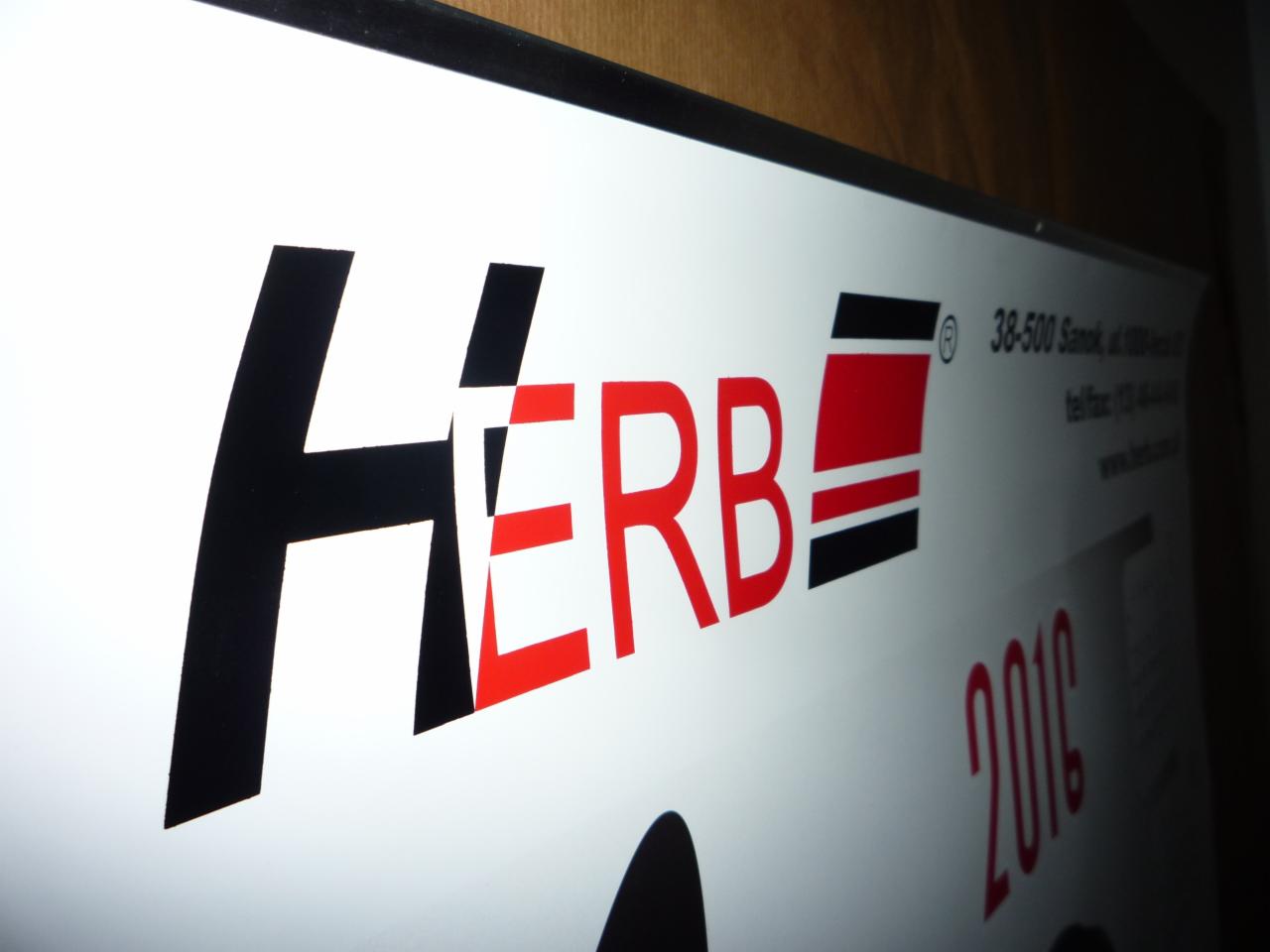 Biała tablica z logo 'Herb' w czerni i czerwieni, adres firmy i rok '2016' w prawym górnym rogu, widoczne pod kątem w oświetlonym pomieszczeniu.