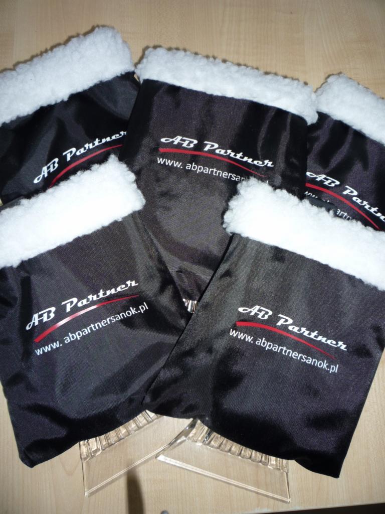 Czarne skrobaczki do szyb z białym futerkiem i nadrukiem 'AB Partner' na przezroczystych podstawkach, leżące na drewnianym blacie. Widok z góry.