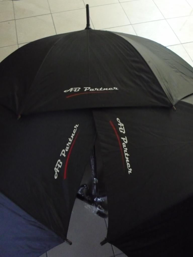 Czarne parasole z nadrukiem 'AB Partner' leżące na kafelkach, widoczne z góry. Nadruk w kolorze białym z czerwonym akcentem. Jeden parasol częściowo złożony.