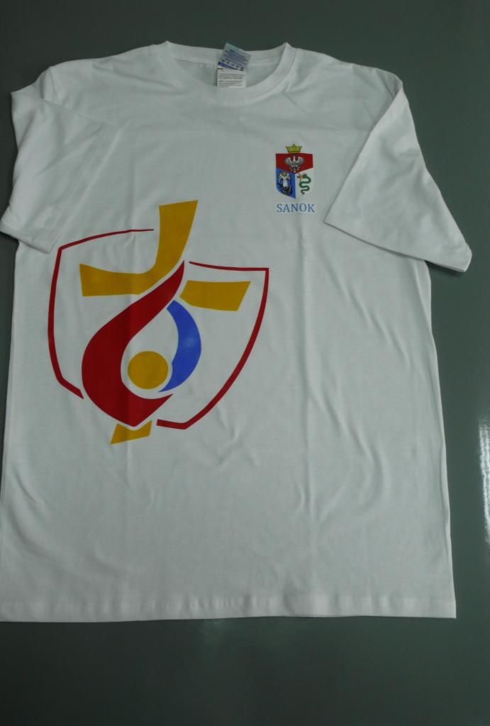 Biała koszulka z nadrukiem herbu Sanoka i stylizowanego krzyża w barwach czerwieni, złota i błękitu. Widoczne logo producenta odzieży. Prezentacja gotowego produktu.