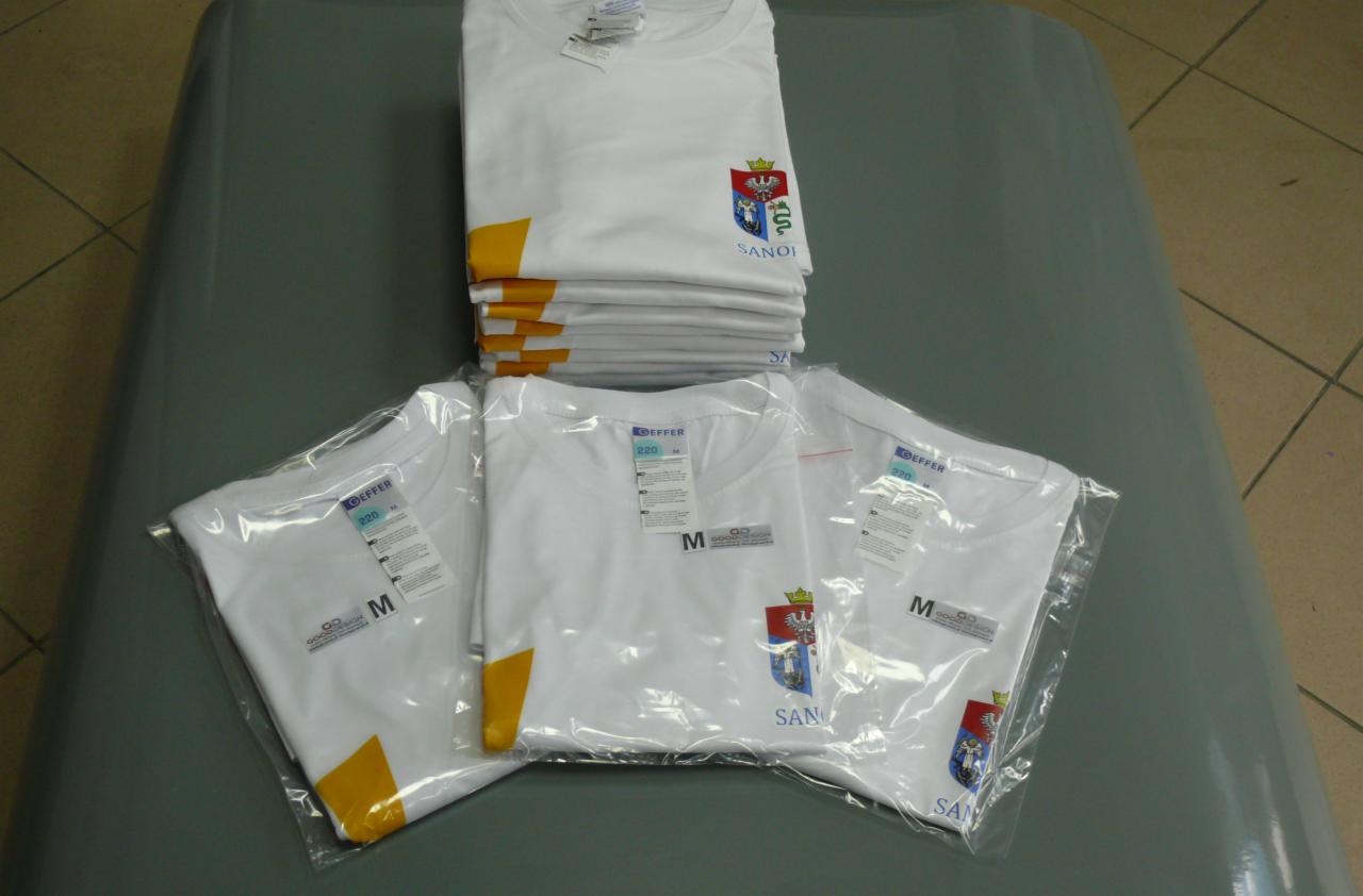 Białe koszulki z nadrukiem herbu i napisu 'Sanok', część w folii, ułożone w stos, widoczne metki i oznaczenia rozmiarów. Nadruk na koszulce.