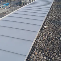 Widok z góry na pomalowany na szaro dach z blachy trapezowej, otoczony kamieniami, z widocznym fragmentem miasta w tle.