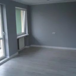 Pomalowane na szaro puste pomieszczenie z panelami podłogowymi, białymi listwami przypodłogowymi, grzejnikiem pod oknem i widokiem na zewnątrz przez balkonowe drzwi.