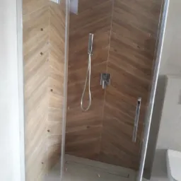 Nowoczesna kabina prysznicowa z drewnopodobnymi płytkami ułożonymi w jodełkę, chromowana armatura, szklane drzwi z minimalistycznym uchwytem, widoczny odpływ liniowy.