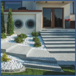 Schody wejściowe z szarej nawierzchni, ozdobione białym grysem i zielenią, prowadzące do domu z oryginalnymi okrągłymi oknami.