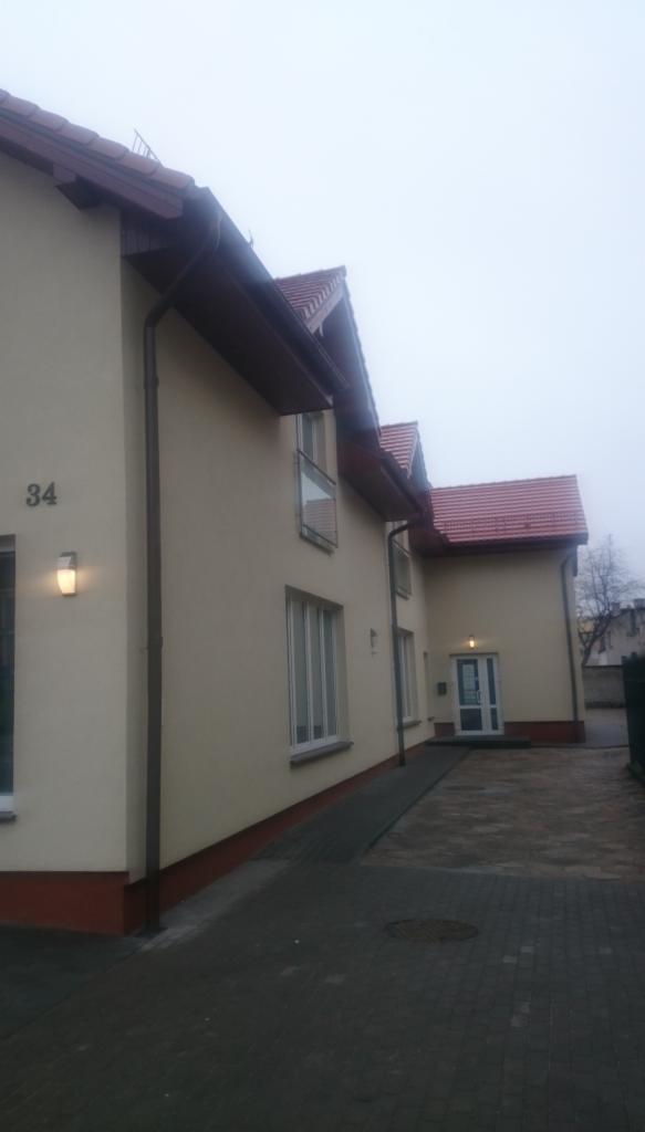 Dwukondygnacyjny dom z beżową elewacją, numerem 34 i czerwonym dachem, sfotografowany w pochmurny dzień z poziomu chodnika.
