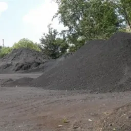 Góra czarnego węgla na placu składowym, widok z dołu, z zielenią drzew w tle. Na drugim planie mniejszy stos. Szare podłoże, pochmurne niebo.