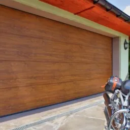 Zamknięta, segmentowa brama garażowa w odcieniach brązu z poziomymi panelami, na tle której stoi motocykl z żółtymi elementami.