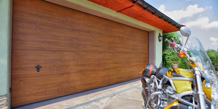 Zamknięta, segmentowa brama garażowa w odcieniach brązu z poziomymi panelami, na tle której stoi motocykl z żółtymi elementami.