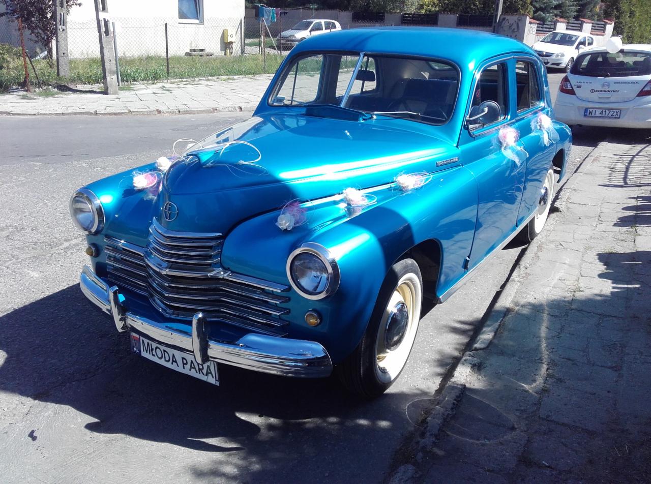 Błękitny, odrestaurowany GAZ-21 Wołga z dekoracjami ślubnymi, tablicą 'Młoda Para' i chromowanymi detalami, zaparkowany na ulicy w słoneczny dzień.
