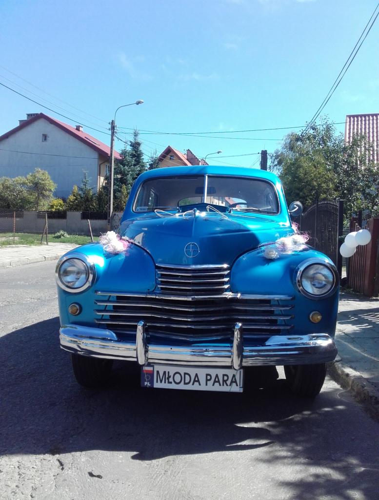 Błękitny, zabytkowy samochód udekorowany kwiatami i tablicą 'Młoda Para' na tle ulicy i domów w słoneczny dzień. Klasyczny pojazd do ślubu.
