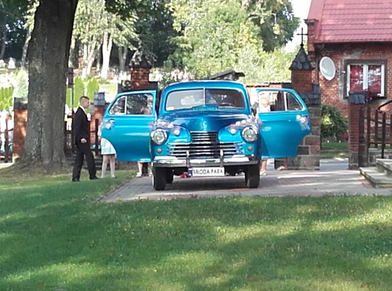 Turkusowy, zabytkowy samochód z otwartymi drzwiami i tablicą 'Młoda Para' na tle zieleni i ceglanego budynku z krzyżem. W pobliżu stoi mężczyzna w garniturze.