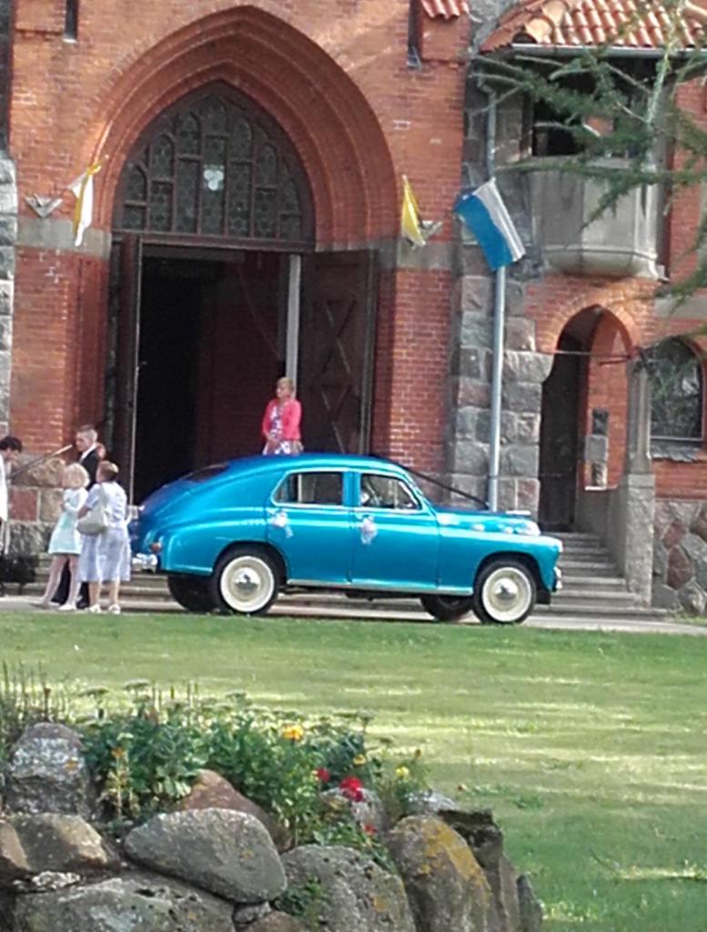 Błękitny, udekorowany kwiatami klasyk przed ceglanym kościołem, goście weselni w tle. Auto z białymi oponami, dekoracje ślubne na karoserii.