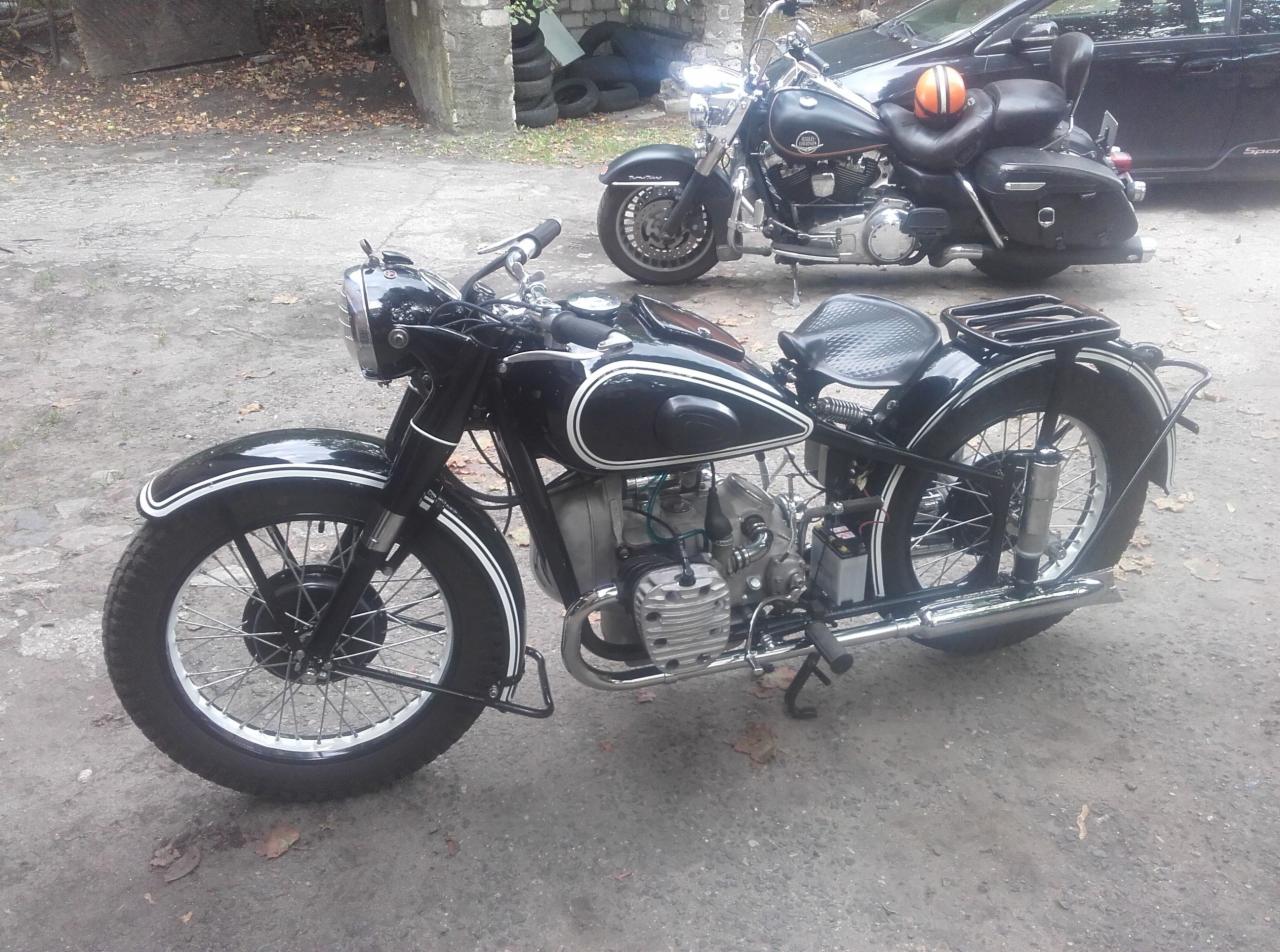 Czarny, klasyczny motocykl Ural z białymi akcentami, zaparkowany na zewnątrz obok innego motocykla i samochodu. Widoczne detale silnika i opony.