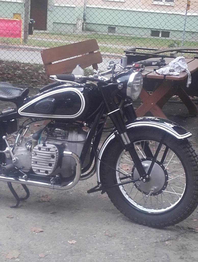 Czarny motocykl z białymi akcentami, widok z boku, zaparkowany na zewnątrz, z drewnianym stołem i narzędziami w tle. Detale vintage, opona z bieżnikiem.