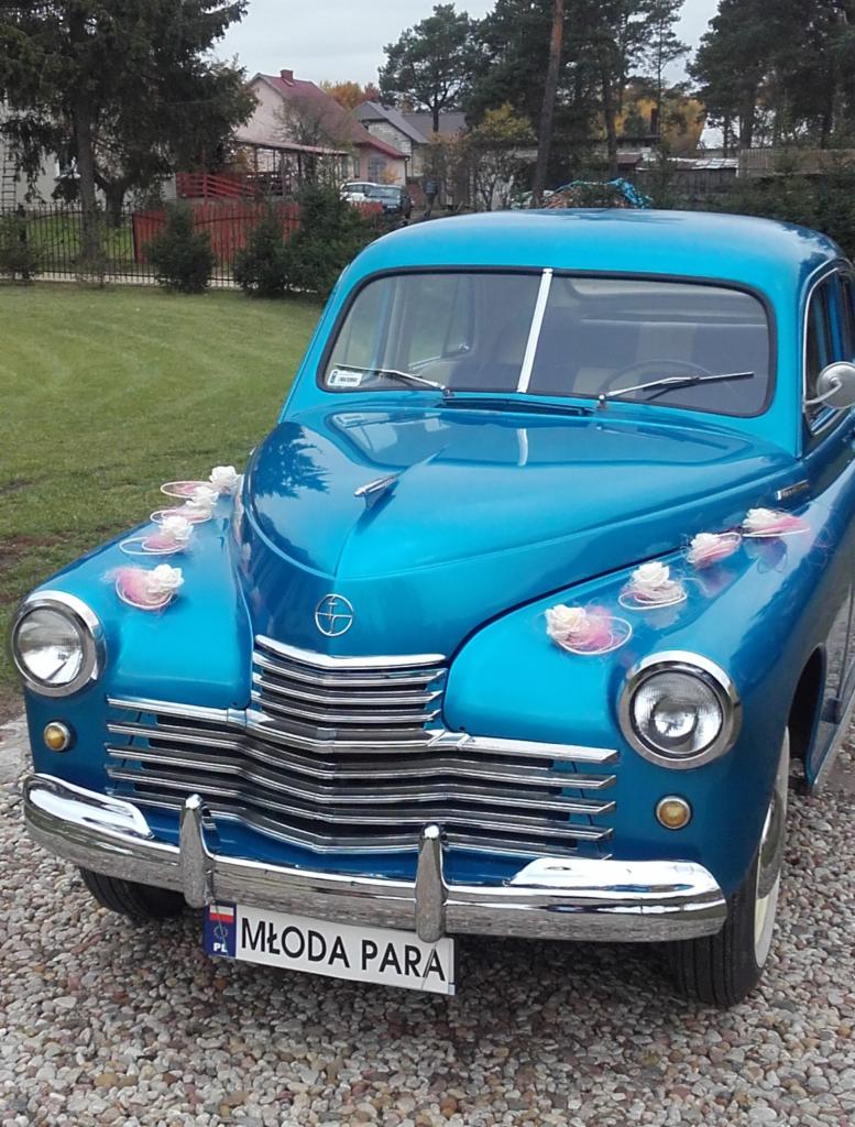 Błękitny, klasyczny samochód udekorowany kwiatami i tablicą 'Młoda Para' na żwirowym podłożu, otoczony zielenią i budynkami w tle.