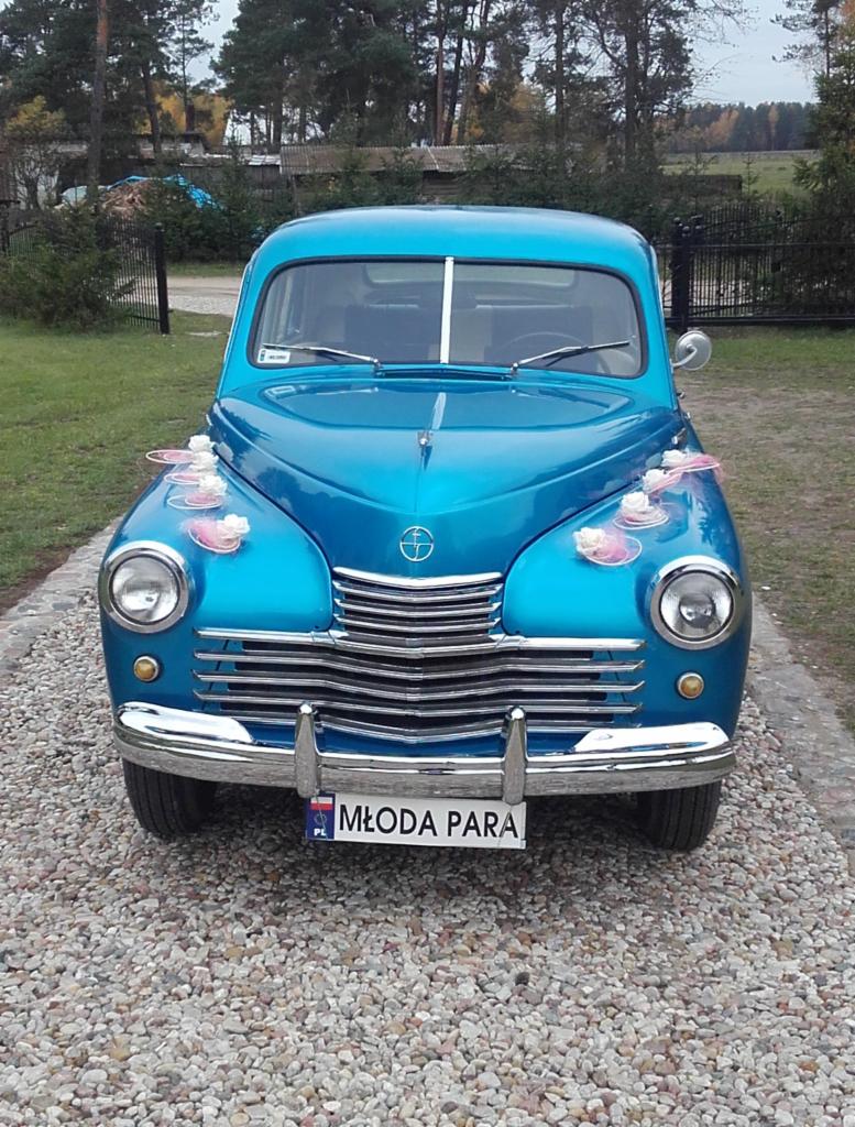 Błękitny, zabytkowy samochód marki GAZ z dekoracjami ślubnymi na masce i tablicą rejestracyjną 'Młoda Para' na tle zieleni i żwirowej drogi.