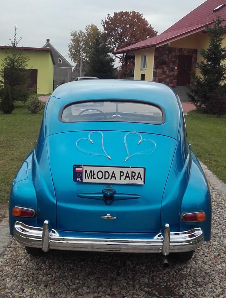 Błękitny, odrestaurowany oldtimer z tablicą 'Młoda Para' i serduszkami, widziany z tyłu na żwirowej drodze, w tle zabudowania. Auto do ślubu.