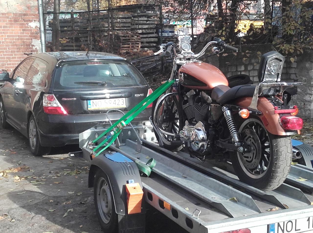 Motocykl w kolorze miedzianym zabezpieczony pasami na platformie przyczepy, holowanej przez czarny samochód osobowy. Widok z tyłu, jesienna sceneria.