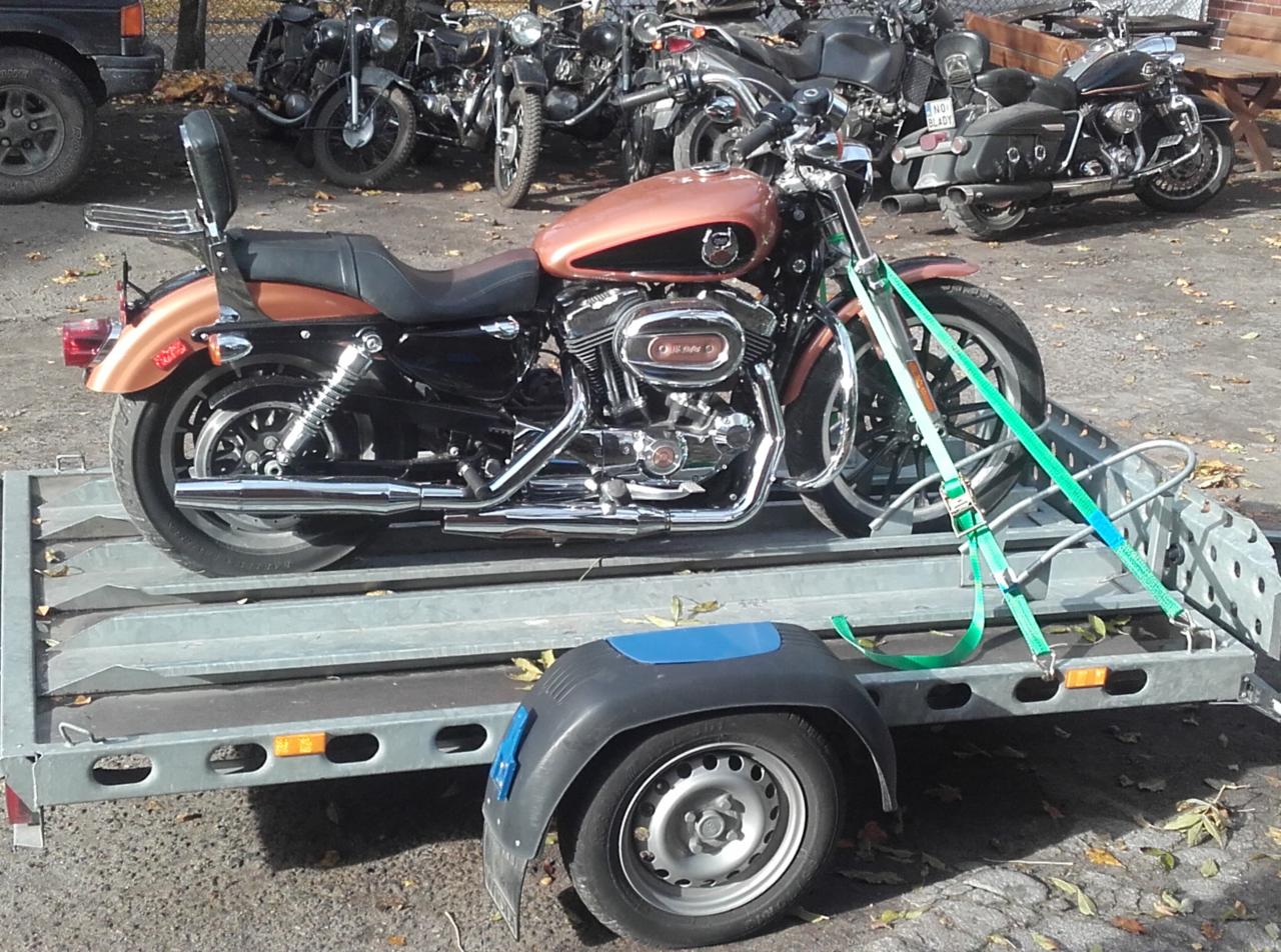Brązowy motocykl Harley-Davidson na szarej przyczepie transportowej, zabezpieczony zielonymi pasami. W tle inne motocykle i fragment samochodu.