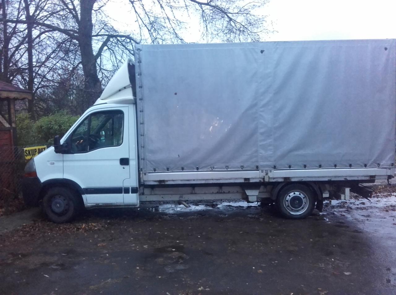 Biały bus z plandeką, widok z boku na mokrym podłożu. Na plandece widoczne ślady użytkowania. Tło stanowią drzewa i zabudowania.