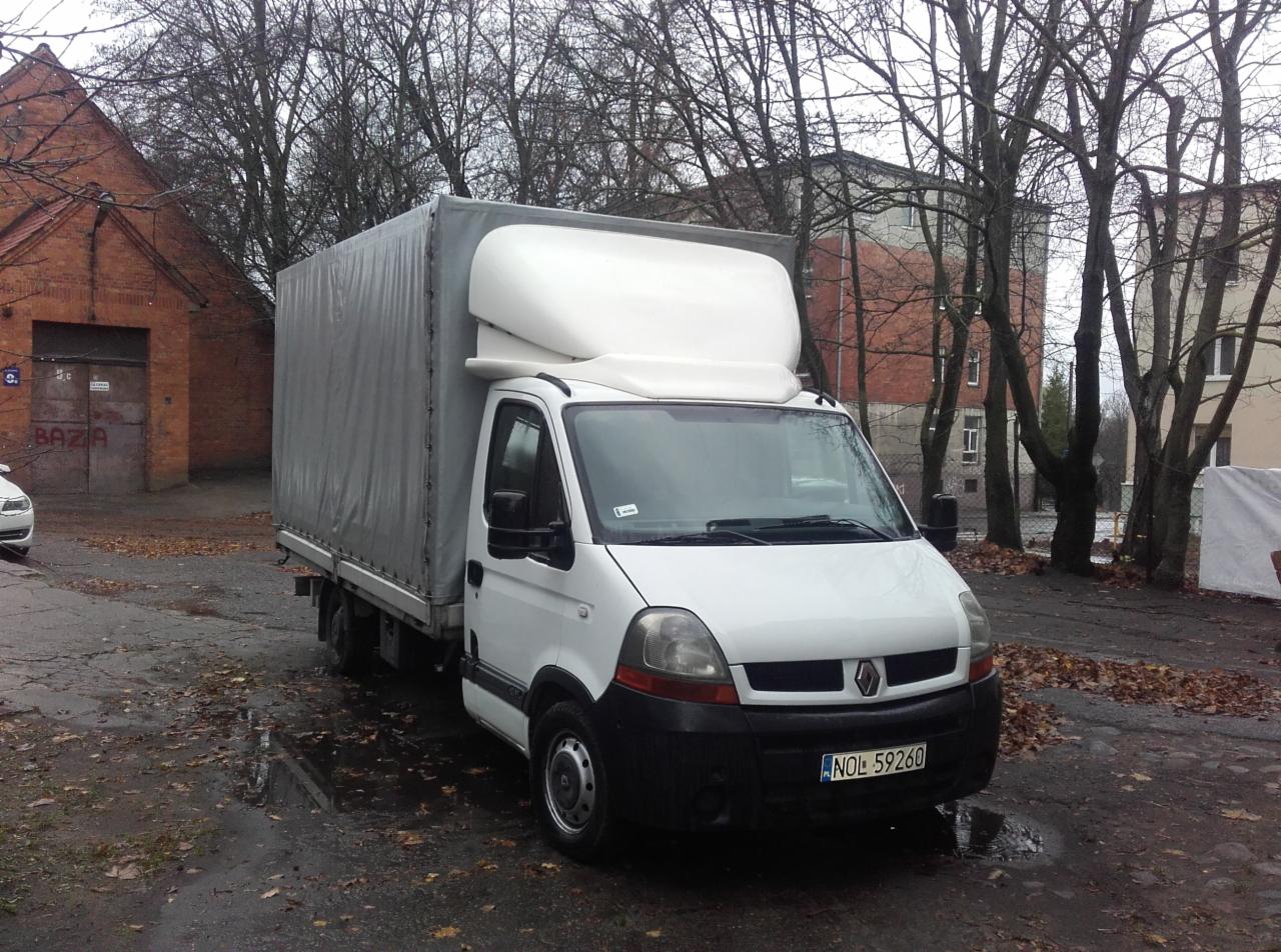 Biały bus Renault Master z plandeką i kabiną sypialną parkuje na tle ceglanego budynku i drzew bez liści. Widoczna tablica rejestracyjna.