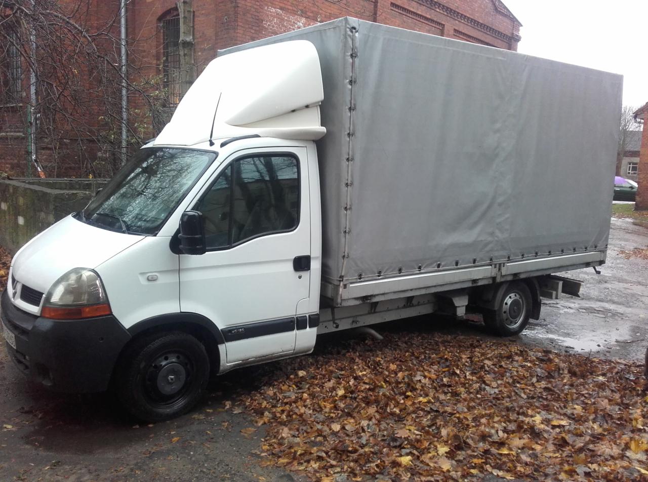Biały bus dostawczy z szarą plandeką na tle ceglanego budynku, zaparkowany na ulicy pokrytej opadłymi liśćmi. Widok z boku, jesienna sceneria.