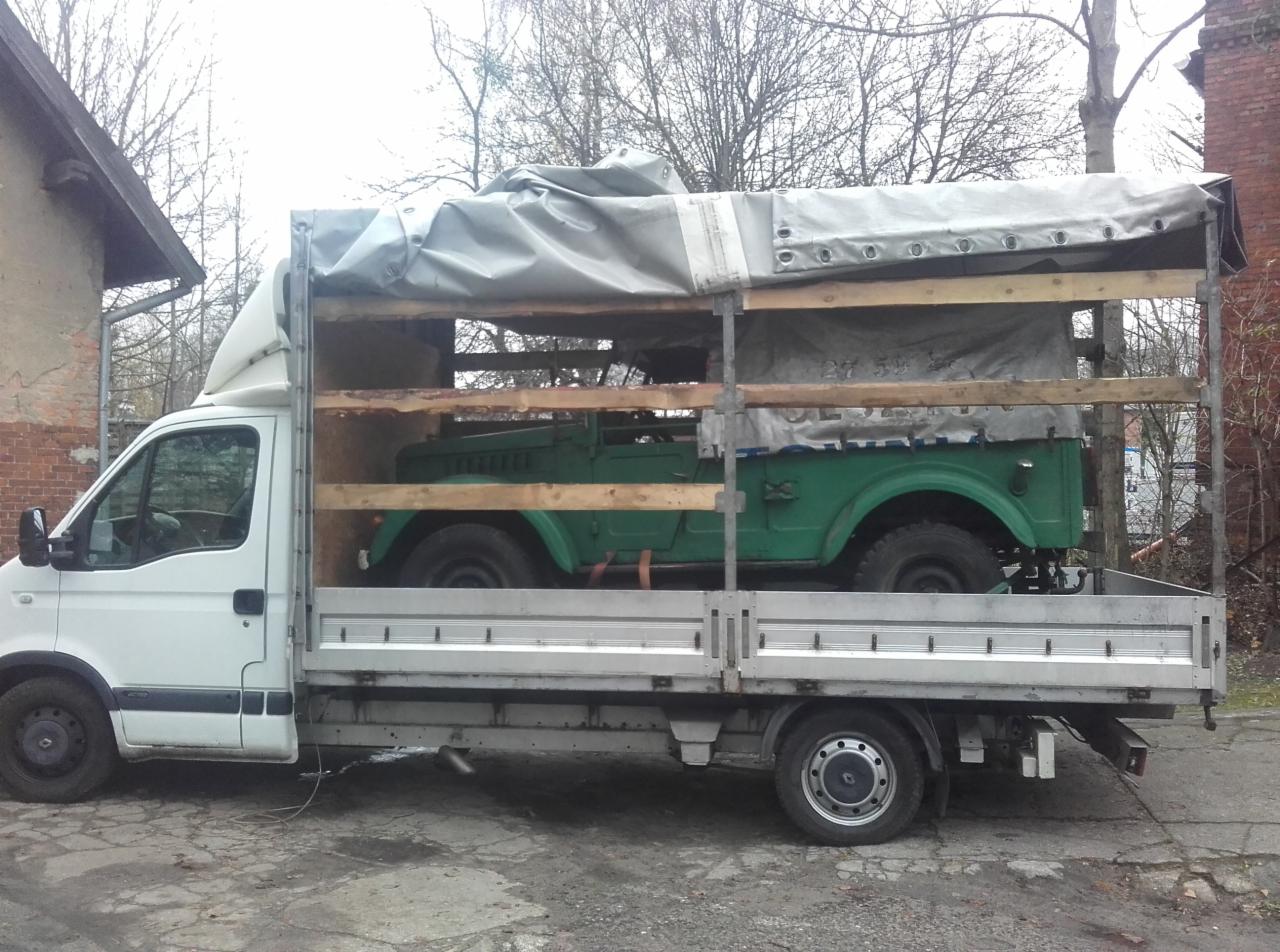 Zielony samochód terenowy zabezpieczony w skrzyni na platformie białego busa dostawczego, przygotowany do transportu. Widok z boku, szare tło.