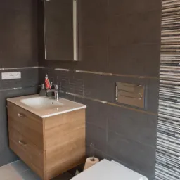 Nowoczesna łazienka w Mońkach z szarymi płytkami, drewnianą szafką pod umywalką i toaletą. Na ścianie dekoracyjna mozaika z pasków.