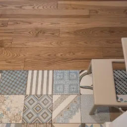 Fragment podłogi w apartamencie w Warszawie: połączenie drewnianych paneli i wzorzystych, patchworkowych płytek ceramicznych w odcieniach brązu i błękitu, z narożnikiem krzesła.