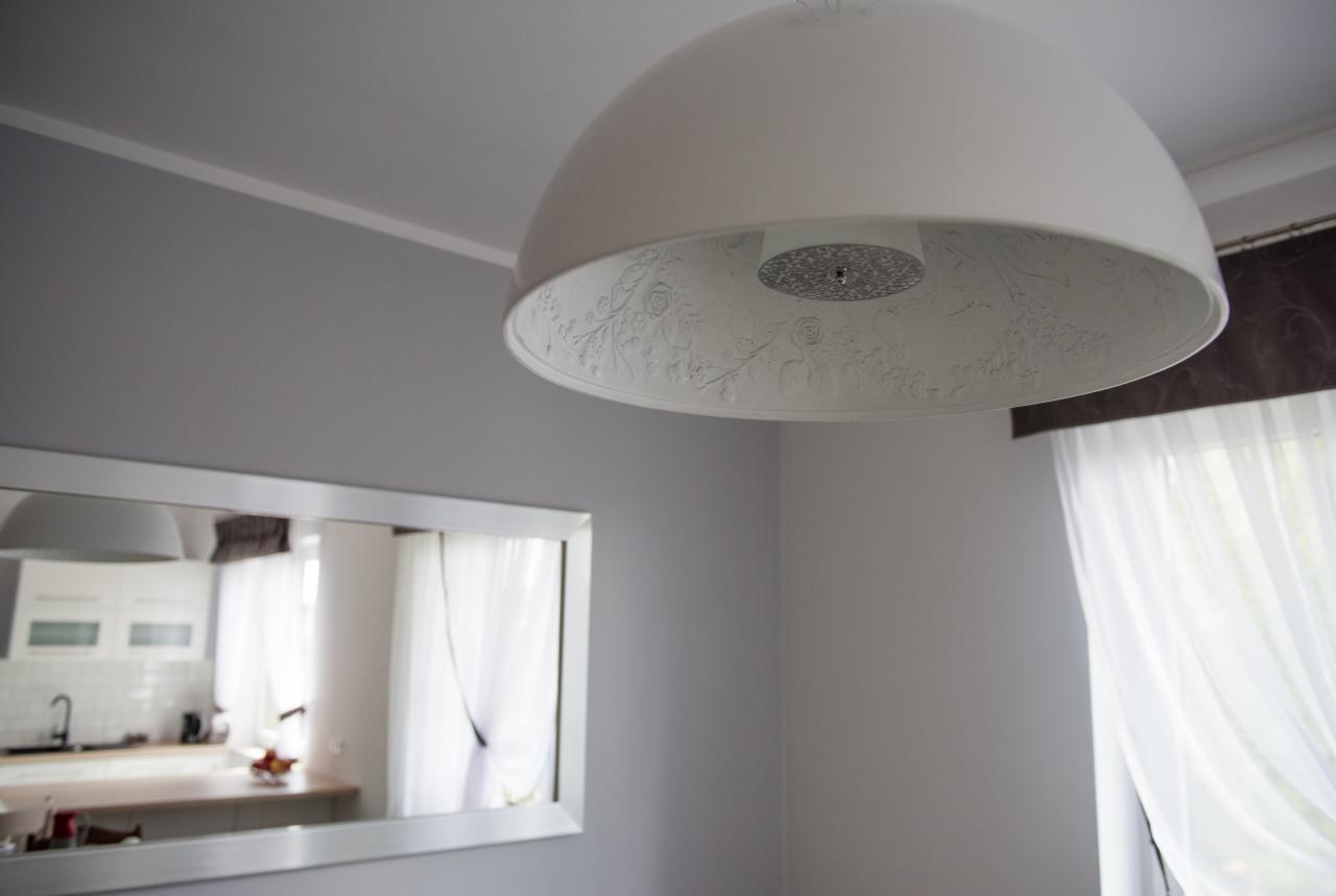 Biała, kopułowa lampa sufitowa z wytłaczanym motywem kwiatowym, odbicie kuchni w lustrze na szarej ścianie.