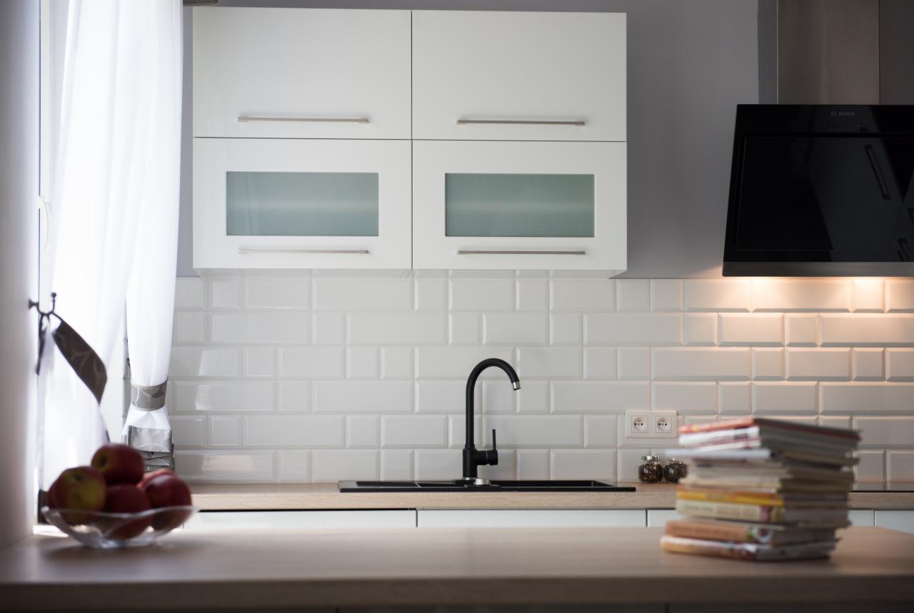 Minimalistyczna kuchnia z białymi, wiszącymi szafkami z matowymi szybami, czarną armaturą i okapem Bosch, uzupełniona stosem książek i misą z jabłkami na drewnianym blacie.