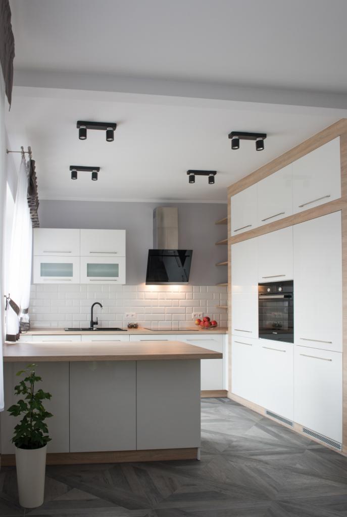 Nowoczesna kuchnia w odcieniach bieli i szarości z drewnianymi akcentami, wyspą kuchenną i podłogą ułożoną w jodełkę. Na blacie widoczne owoce w misce.