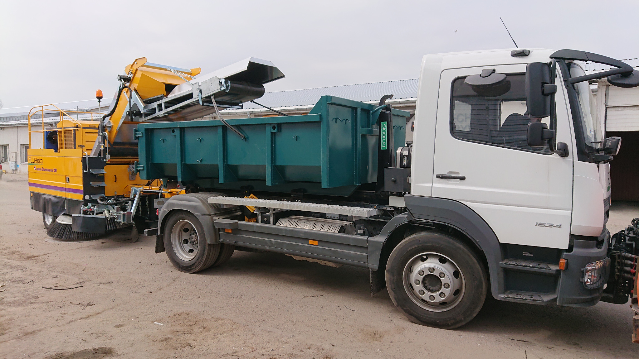 Żółto-fioletowa zamiatarka FlorEko podłączona do ciężarówki z zielonym kontenerem na odpady, widoczne szczotki i mechanizm podający materiał do kontenera.