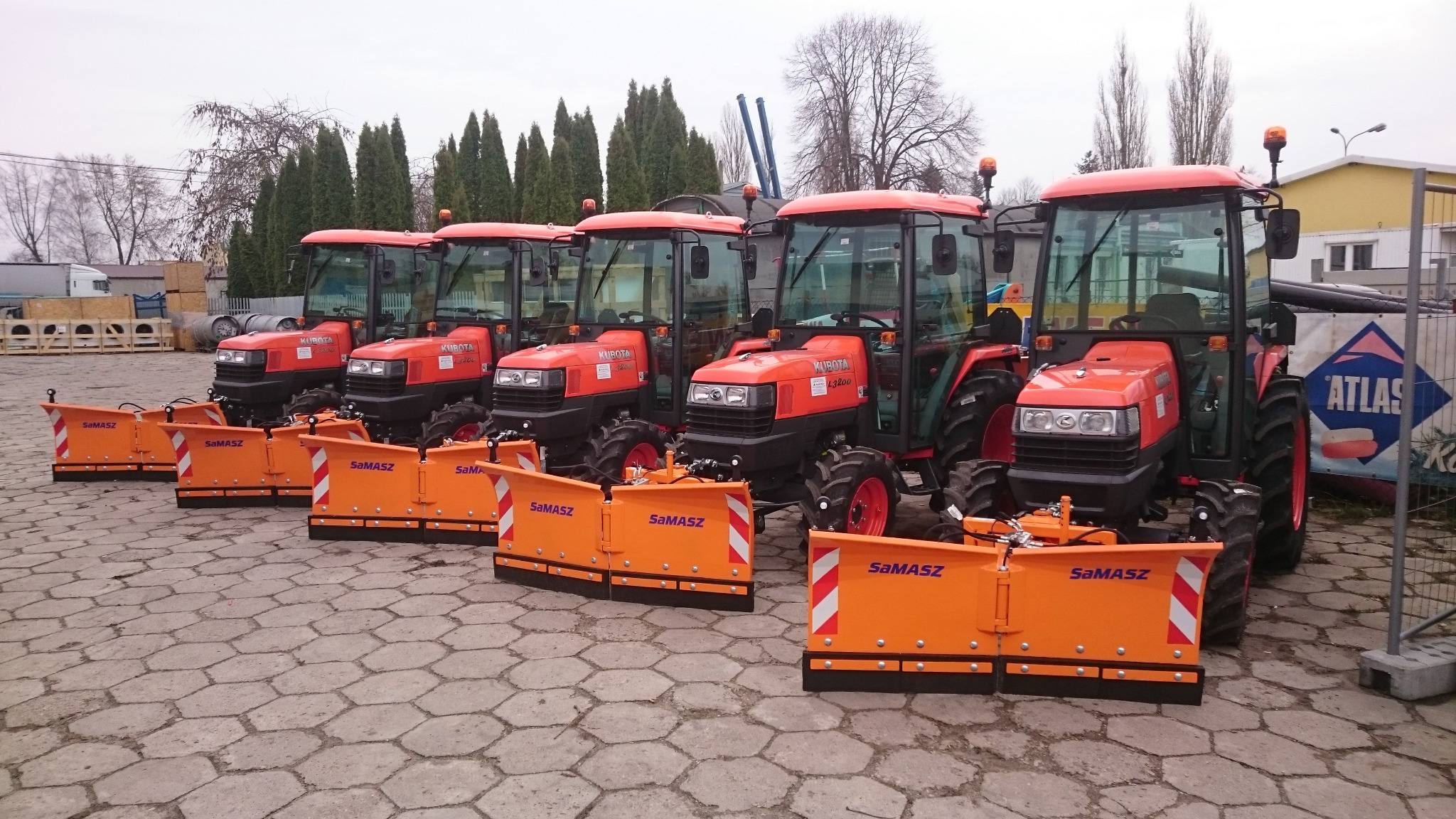 Rząd pomarańczowych traktorów Kubota z pługami śnieżnymi SaMASZ, ustawionych na brukowanym placu, z drzewami iglastymi w tle.