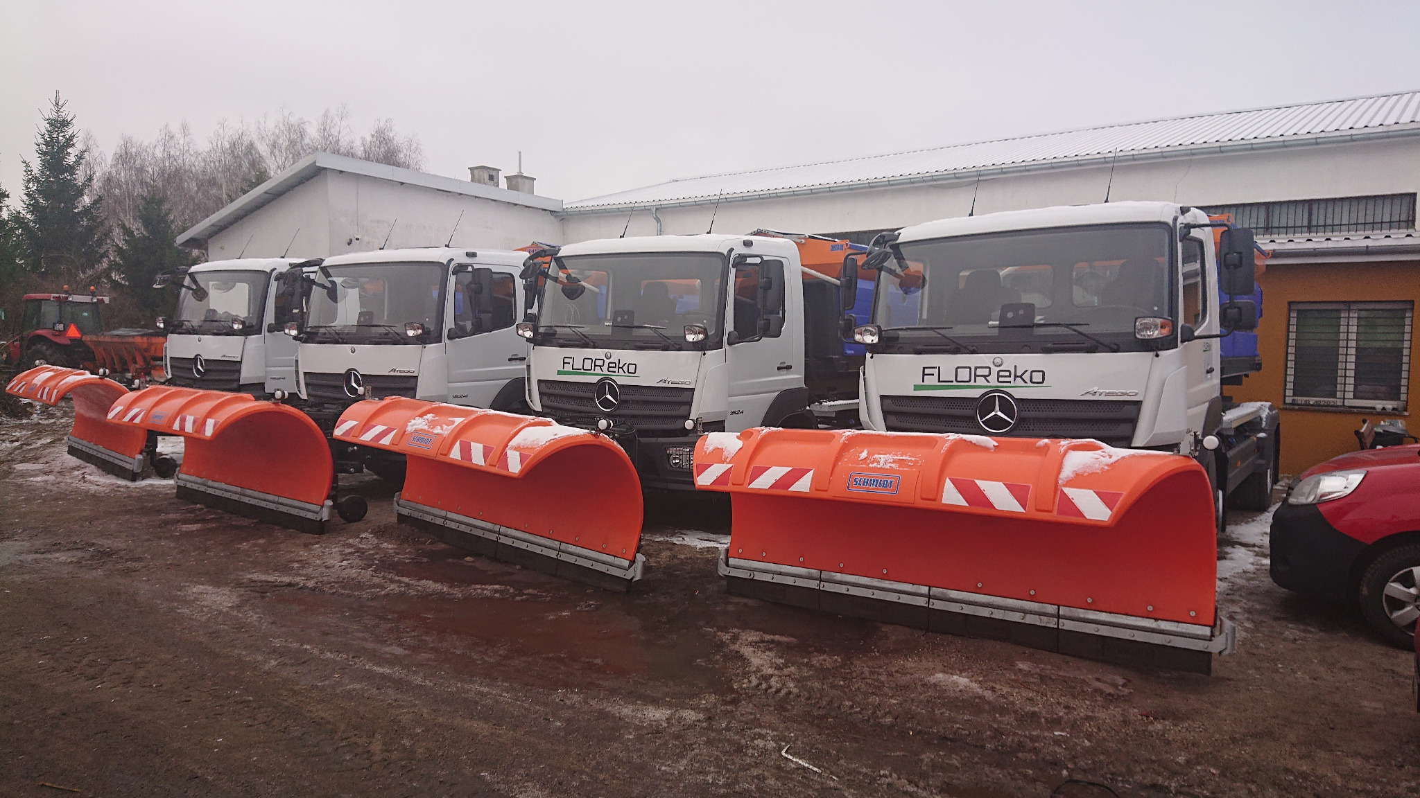 Flota białych ciężarówek z pomarańczowymi pługami śnieżnymi marki Schmidt, zaparkowana na błotnistym placu w pochmurny dzień. Na budynkach w tle widoczny śnieg.