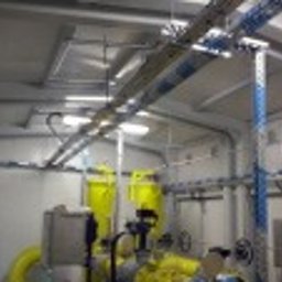 E-funia Projekty techniczne, Automatyka przem., Elektryka, kompensacja mocy biernej, Falownikowe Układy Napędowe - Wnętrze techniczne z rzędami metalowych korytek kablowych pod sufitem, żółte rury i zawory. Widok na instalacje w pomieszczeniu z białymi ścianami i sufitem.