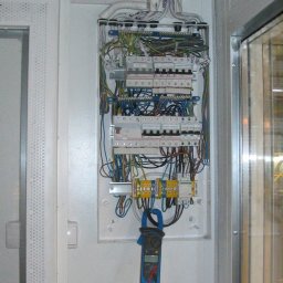 E-funia - Otwarta skrzynka elektryczna z bezpiecznikami i przewodami, pomiar prądu cęgowym miernikiem. Widoczne kable w peszlach. Białe ściany i drzwi.