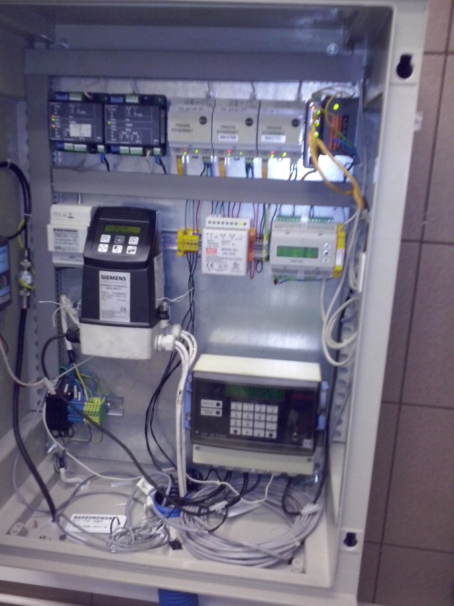 Otwarta szafa sterownicza z widocznymi modułami, przewodami i urządzeniami marki Siemens. Widoczne elementy automatyki przemysłowej i okablowanie wewnątrz obudowy.