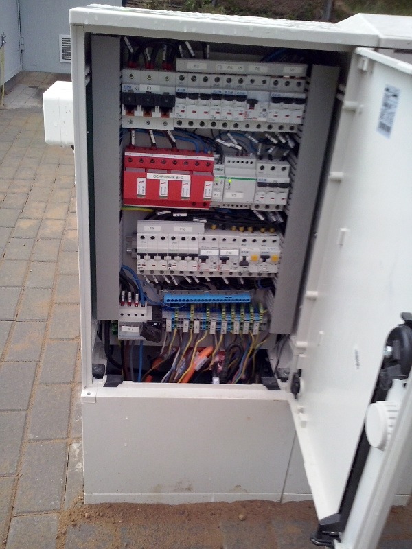 Otwarta skrzynka elektryczna z widocznymi bezpiecznikami, przewodami i elementami instalacji. Widoczne zabezpieczenia przepięciowe. Instalacja w białej obudowie.