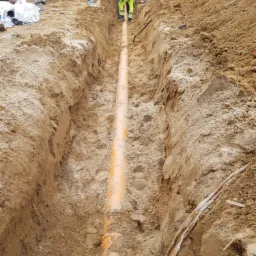 Wykopany rów z ułożoną pomarańczową rurą kanalizacyjną, widoczni pracownicy i koparka w tle, ziemia piaszczysta.