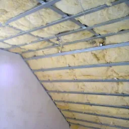 Ocieplenie poddasza wełną mineralną, widoczna metalowa konstrukcja sufitu podwieszanego, skośna ściana. W rogu żółta rolka wełny w folii.