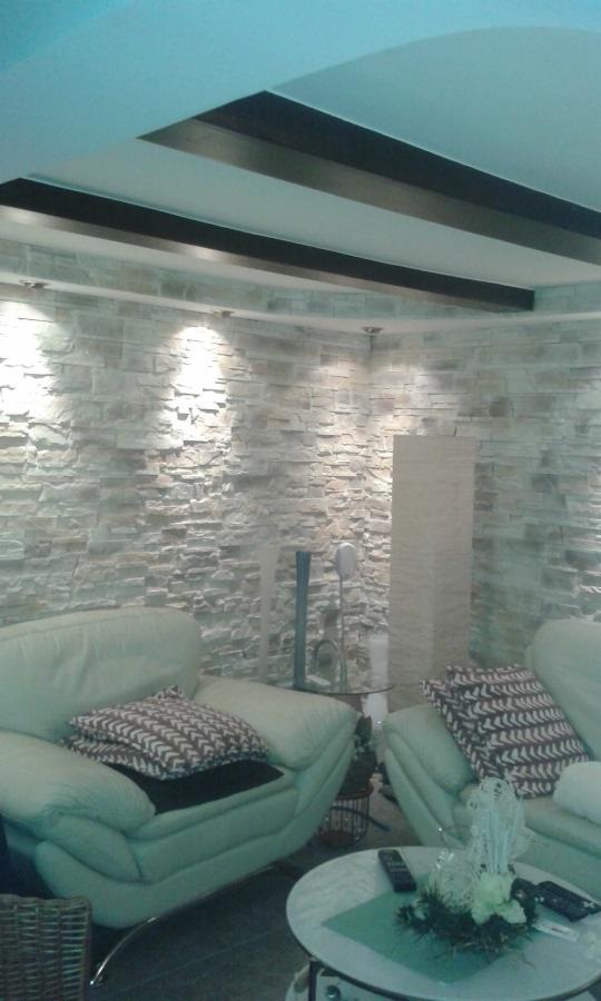 Ściana wykończona dekoracyjnym kamieniem w salonie z oświetleniem punktowym i elementami dekoracyjnymi. Widoczne dwa fotele i stolik kawowy.