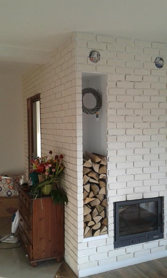 Biała ściana z cegły z wnęką na drewno kominkowe i wkładem kominkowym, obok komoda z kwiatami i elementami dekoracyjnymi.