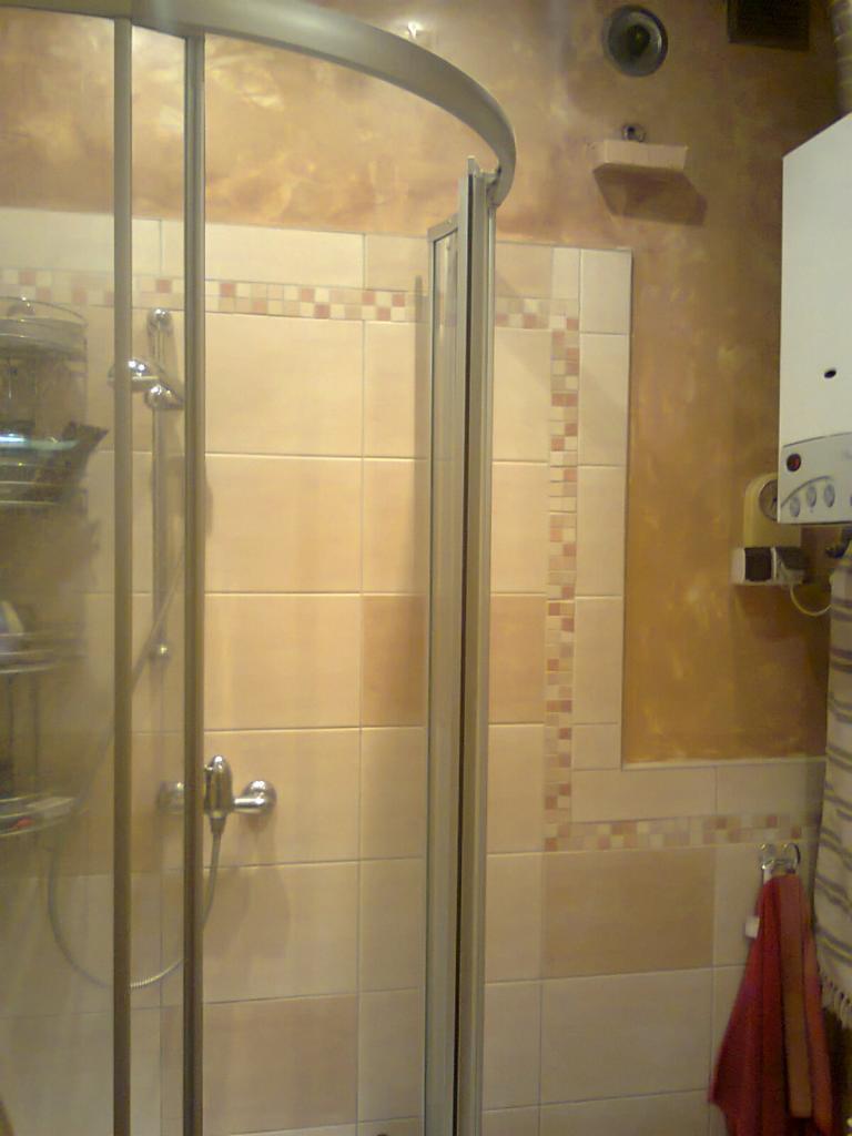 Beżowa łazienka z kabiną prysznicową i dekoracyjnymi paskami z mozaiki. Na ścianie widoczny piec gazowy i ręcznik.