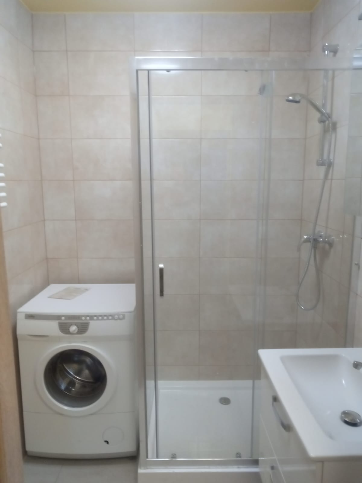 Wykończona łazienka z beżowymi płytkami, kabiną prysznicową z przezroczystymi drzwiami, białą pralką i umywalką z szafką.