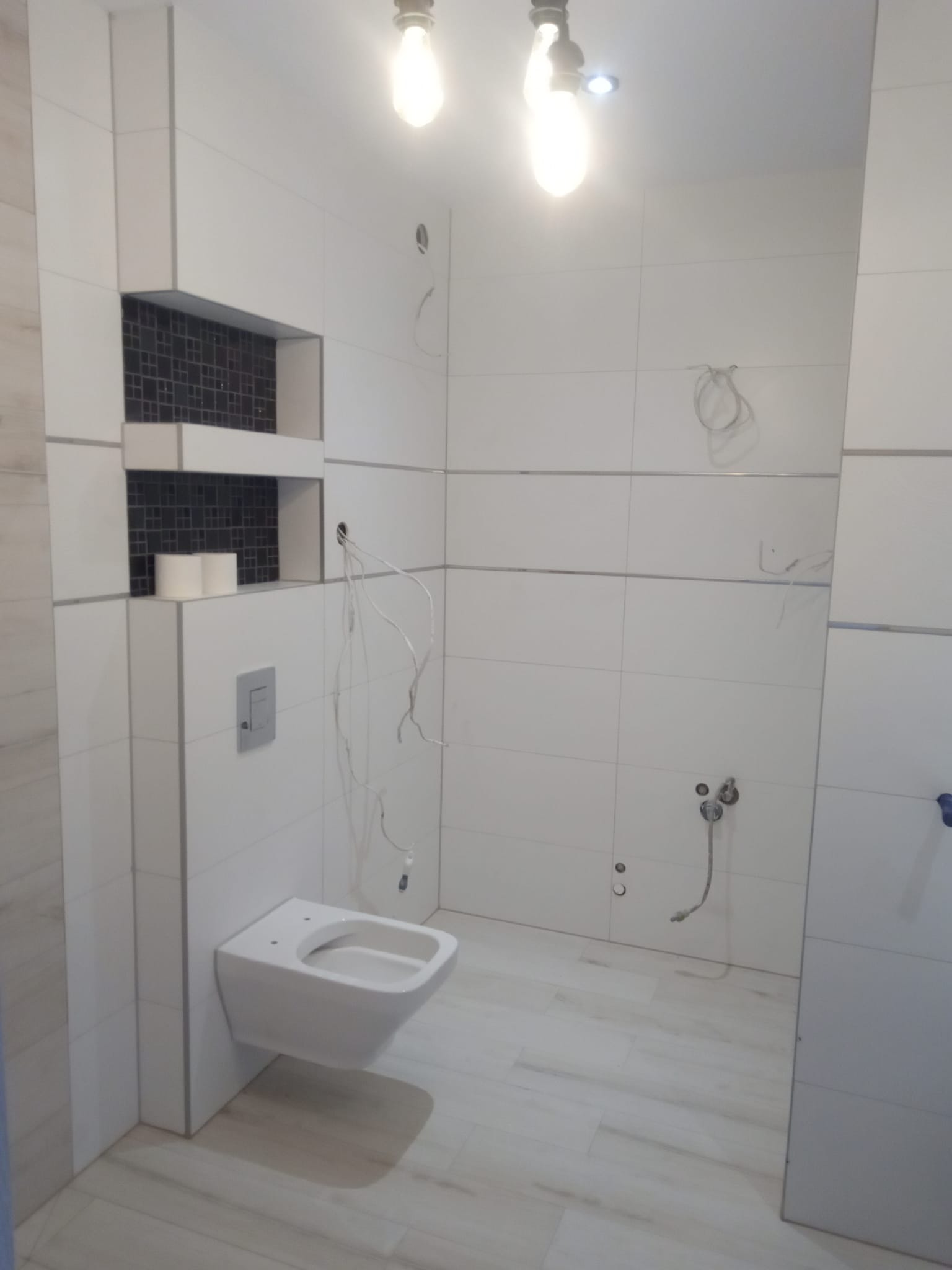 Wnętrze łazienki w trakcie prac wykończeniowych: białe płytki na ścianach i podłodze, podwieszana toaleta, wnęka wyłożona czarną mozaiką, widoczne przewody elektryczne i rury hydrauliczne.