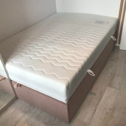 Design 4 Beds Sp. zo.o. - Łóżko jednoosobowe z jasnym materacem i tapicerowaną bazą w odcieniu pudrowego różu, widoczne uchwyty do podnoszenia materaca, częściowo odbite w lustrze.