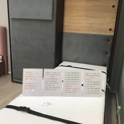 Design 4 Beds Sp. zo.o. - Prezentacja łóżka chowanego w szafie z tapicerowanym panelem, obok materac z czarnym pasem mocującym; widoczne tabliczki informacyjne z cenami i wymiarami.