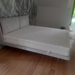 Design 4 Beds Sp. zo.o. - Szare łóżko z tapicerowanym zagłówkiem, ustawione w pokoju z drewnianą podłogą i tapetą w liście; widoczna osoba w tle.